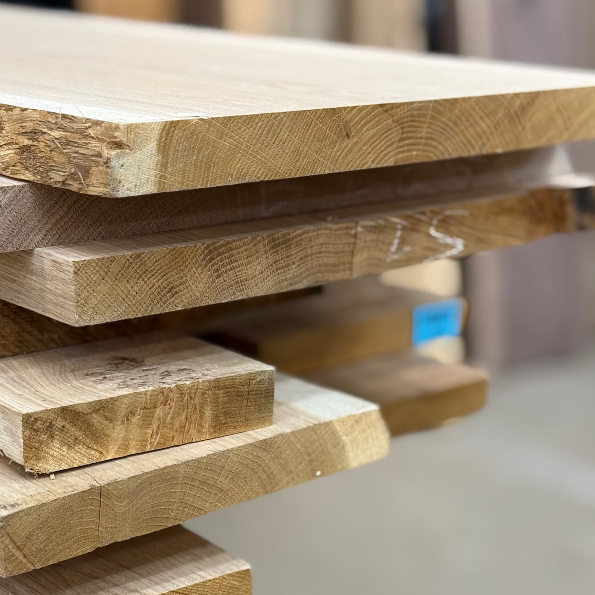 A close-up of stacked natural wood boards and planks with visible grain, photographed in a workshop setting with a blurred background.
