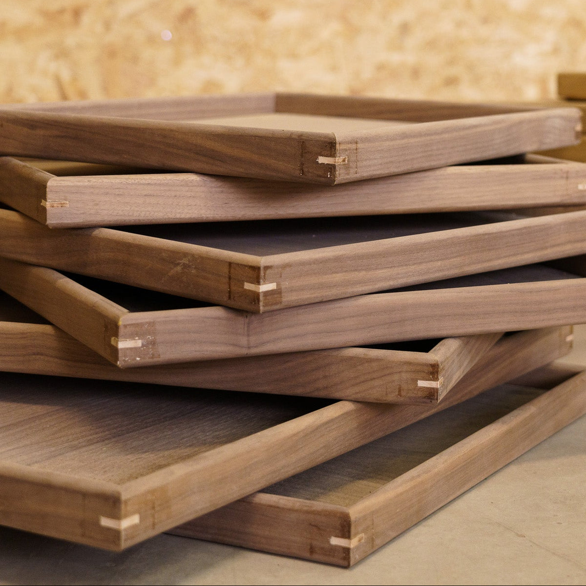Stacks of wooden trays with visible contrasting corner pin details, arranged on a floor against a wood OSB wall.