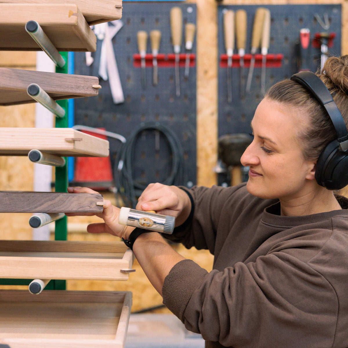 A woman wearing black over-ear headphones works in a woodworking shop, using a small handheld hammer on a wooden tray on a green rack.