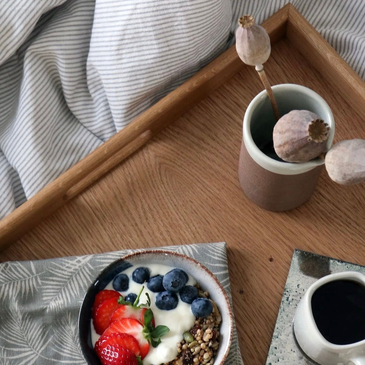 A styled breakfast scene on a wooden tray with a bowl of yogurt topped with strawberries, blueberries, and granola, alongside a ceramic mug and soft bedding in the background.