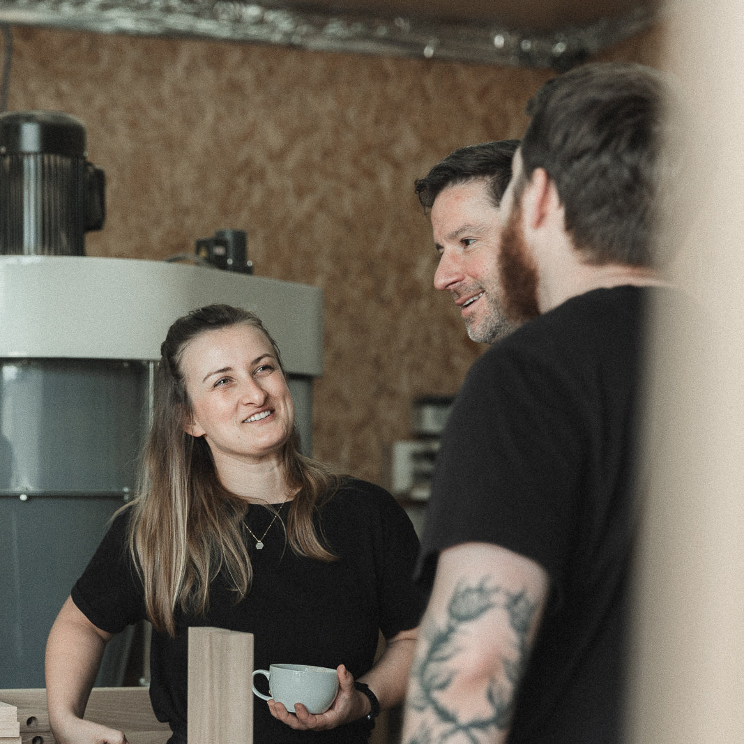 A woman in a black top holding a light blue mug smiles while standing in a workshop next to a wooden workbench, with two men in the foreground in a casual, indoor setting.