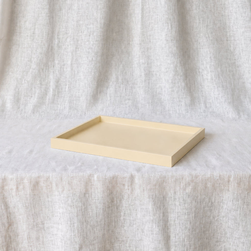 A minimalist rectangular tray in a light cream or pale yellow color, set against a draped, textured off-white fabric backdrop.
