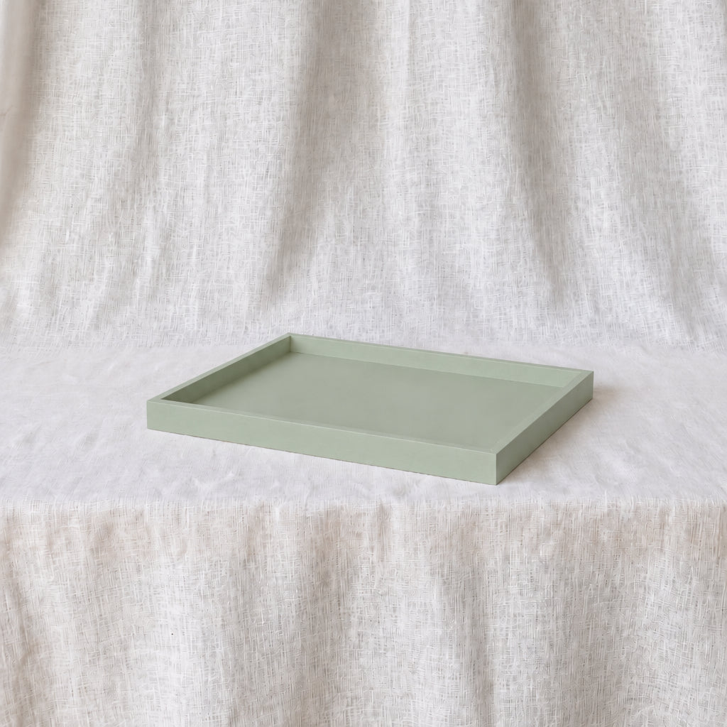 A rectangular sage green tray with raised edges, placed on a textured white fabric surface against a matching draped fabric backdrop.