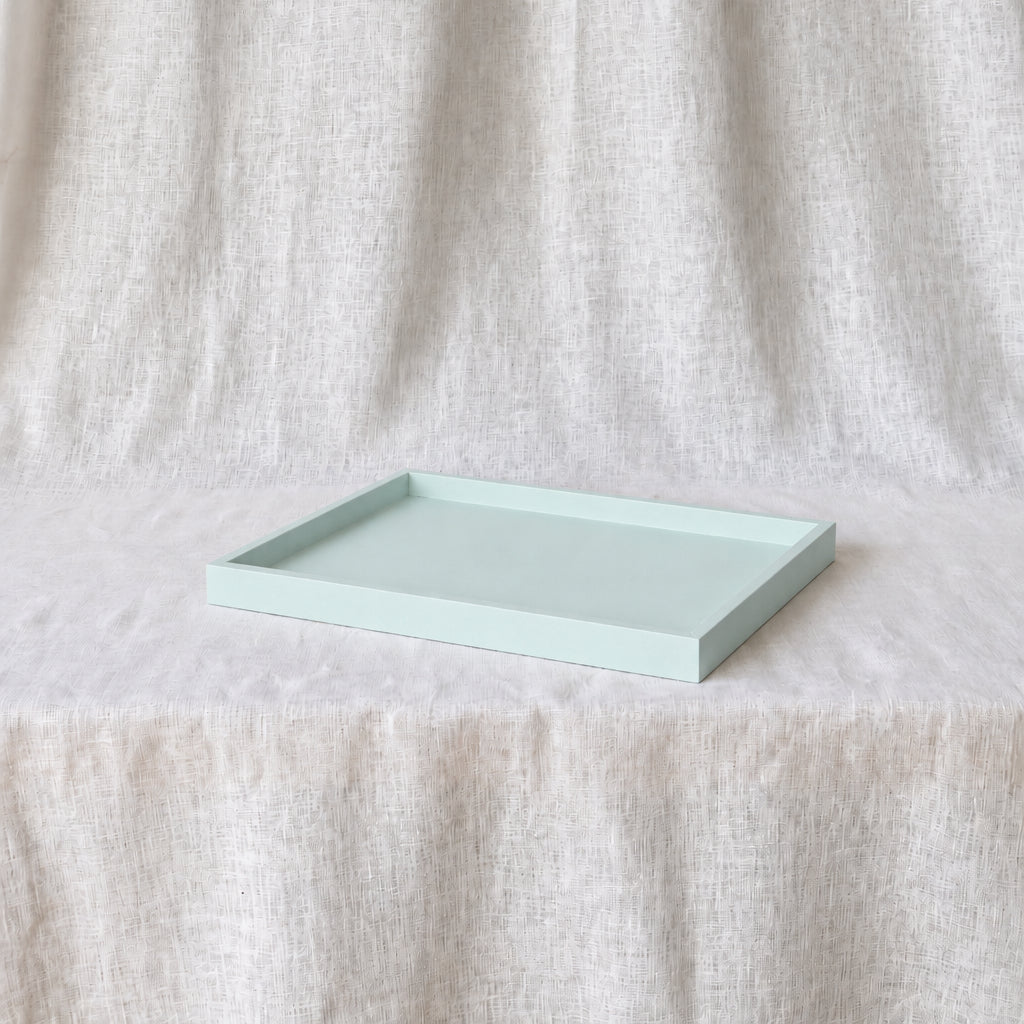 A rectangular mint green tray with a shallow depth and raised edges, resting on a draped cream-colored fabric surface against a matching fabric backdrop.