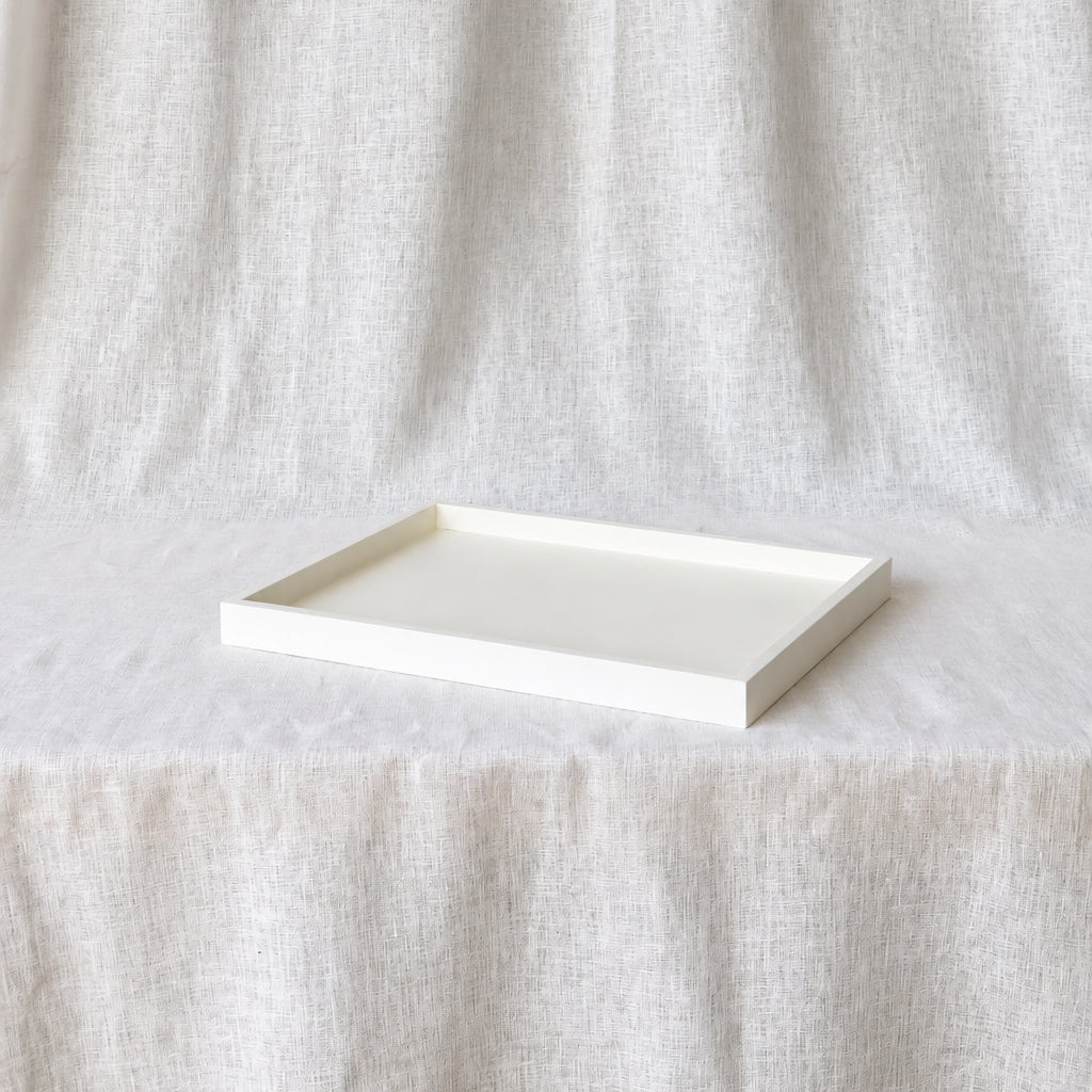 A minimalist rectangular tray with a raised rim, shown in a warm off-white or cream color, resting on a draped linen fabric backdrop.