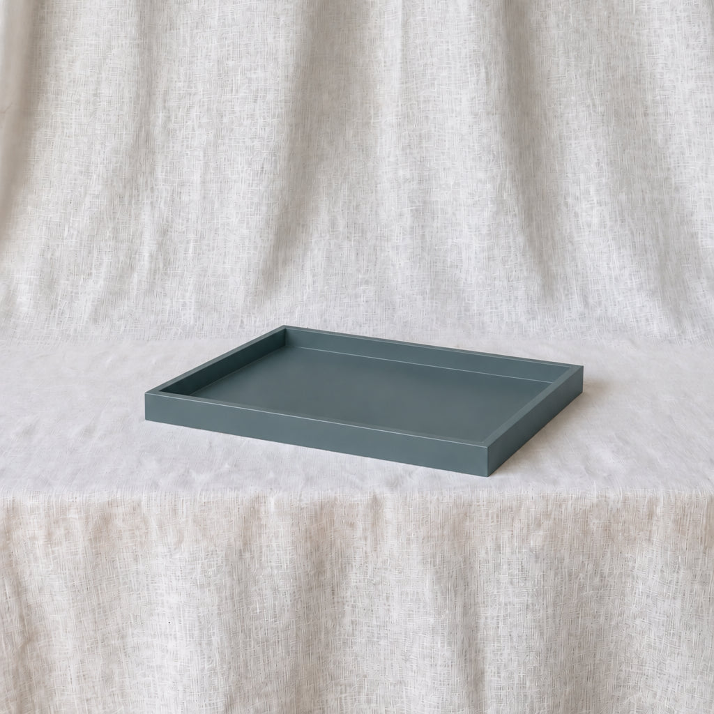 A rectangular matte smoke-blue tray with raised edges, placed on a draped light beige fabric surface against a matching fabric backdrop.