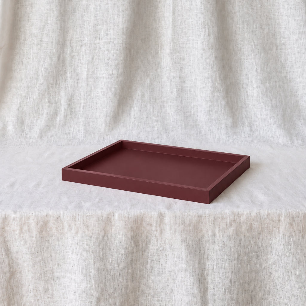 A rectangular matte tray in a deep burgundy red color, placed on a white textured fabric surface with a matching draped fabric backdrop.