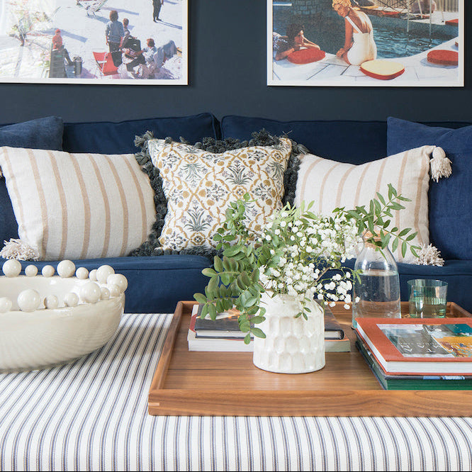 A styled living room scene with a navy blue sofa, striped and patterned throw pillows, two framed art prints hanging on a dark navy wall, a striped upholstered footstool and a medium brown walnut tray holding books and a white vase with greenery.