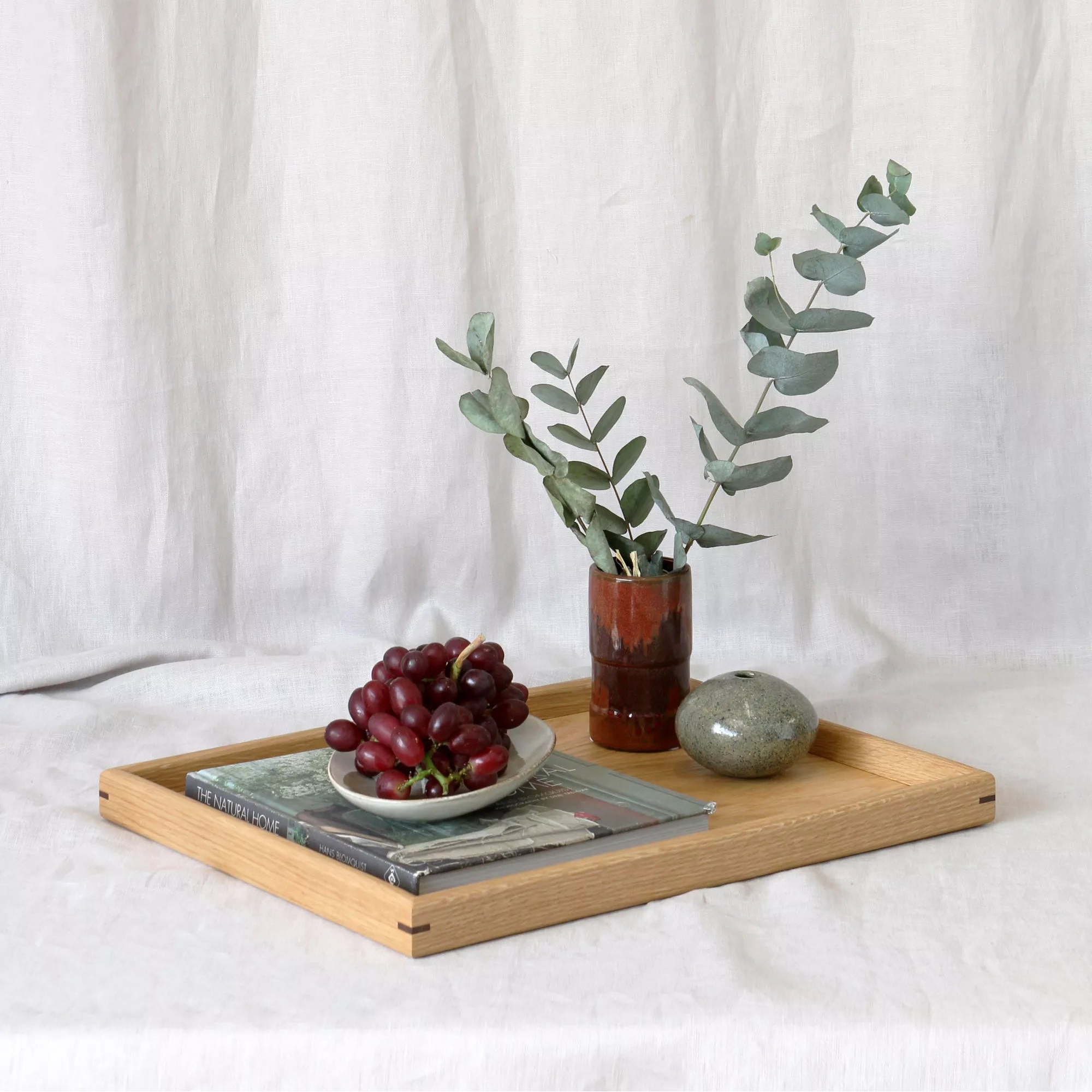 Wooden oak tray with grapes, a vase of eucalyptus on a white fabric background. A large coffee table tray, featuring fine joinery details and a natural oak finish, is designed for functional elegance.