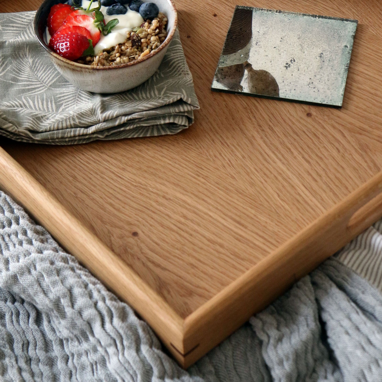Close-up of a handcrafted wooden tray, showcasing a bowl of yogurt with granola and fresh berries, set on a soft gray blanket.