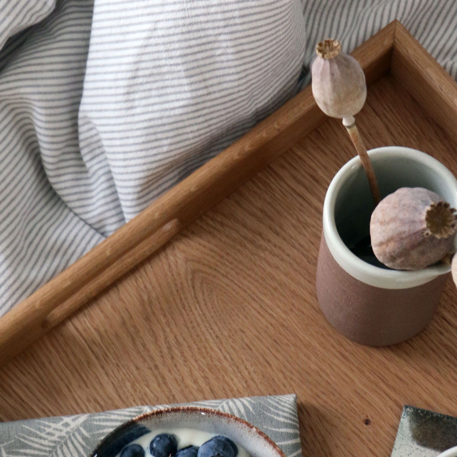 Top-down angle of a wooden tray on a bed, displaying a ceramic vase with dried flowers and a cup of coffee.