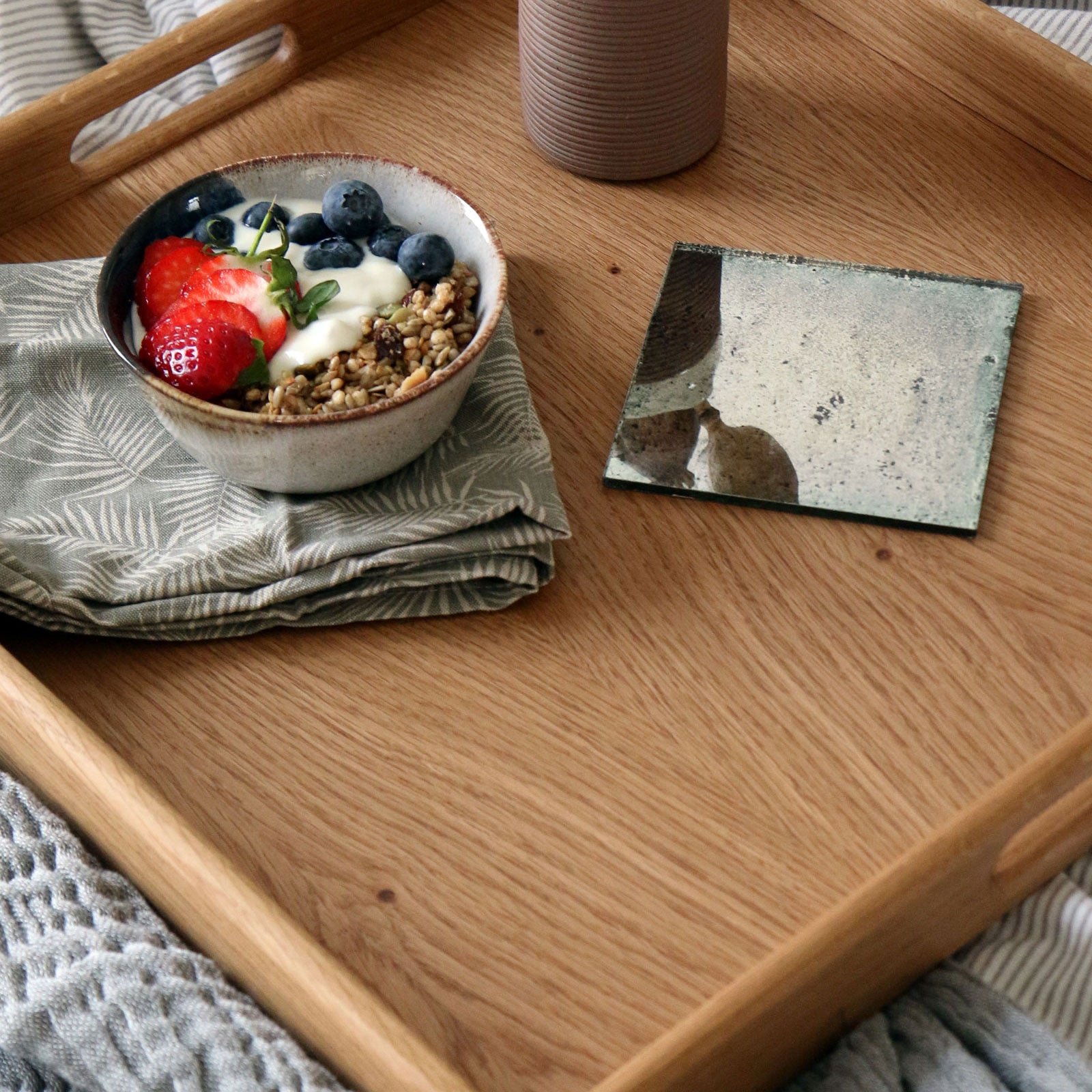 Overhead view of a wooden ottoman tray, holding a breakfast bowl, a textured napkin, and a decorative mirror coaster.