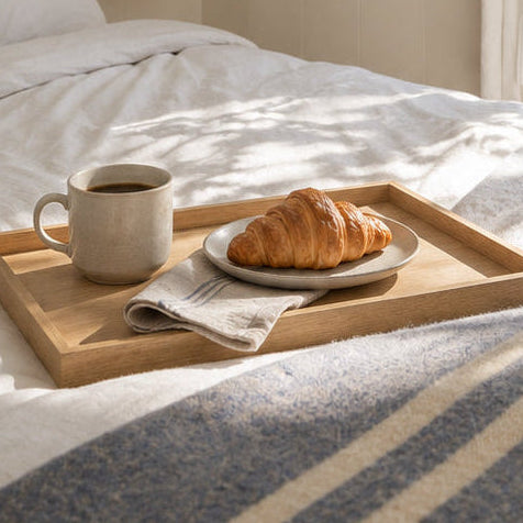 A cozy bedroom scene with a neatly made bed, a wooden tray holding a mug and croissant on the bed, a white robe hanging on a wall hook, and a glass door opening to a sunlit outdoor area with trees.