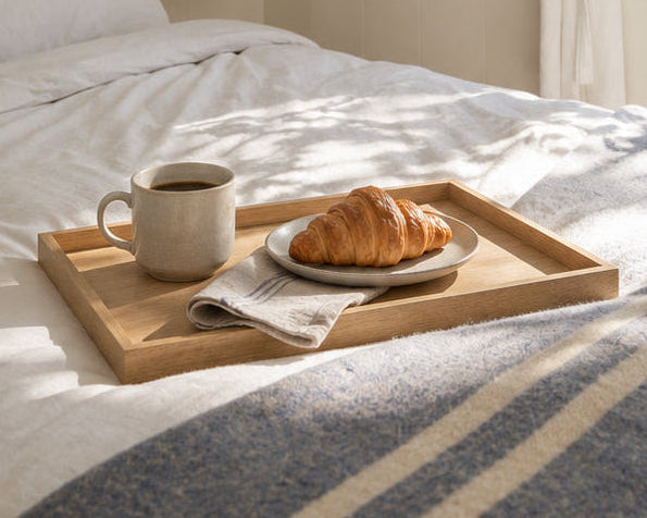 A cozy bedroom scene with a neatly made bed, a wooden tray holding a mug and croissant on the bed, a white robe hanging on a wall hook, and a glass door opening to a sunlit outdoor area with trees.
