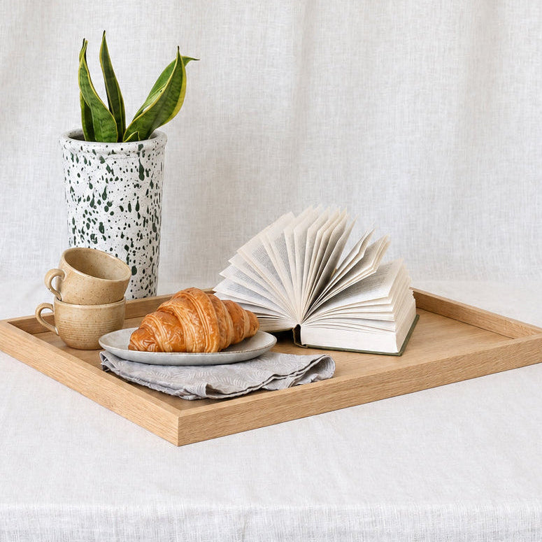 A light wood rectangular tray styled with a ceramic planter, two beige cups, a croissant on a plate, and an open book, set on a white linen-covered table against a matching fabric backdrop.