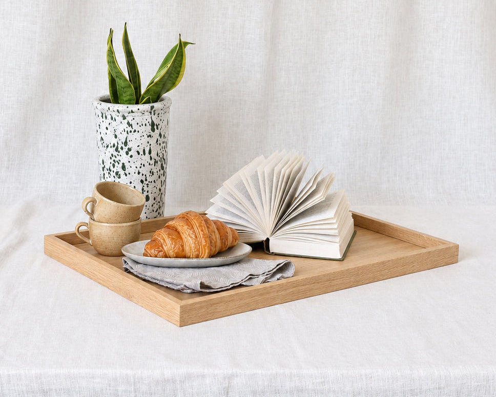 A light wood rectangular tray styled with a ceramic planter, two beige cups, a croissant on a plate, and an open book, set on a white linen-covered table against a matching fabric backdrop.