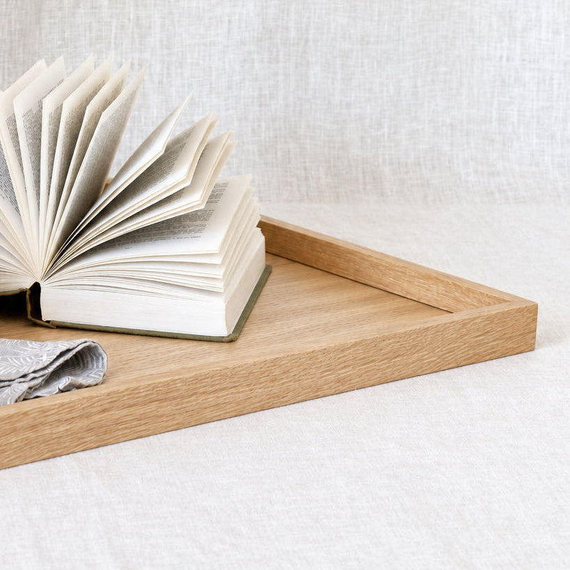 A light natural wood tray holding an open book and a folded gray cloth, set against a soft white linen backdrop.