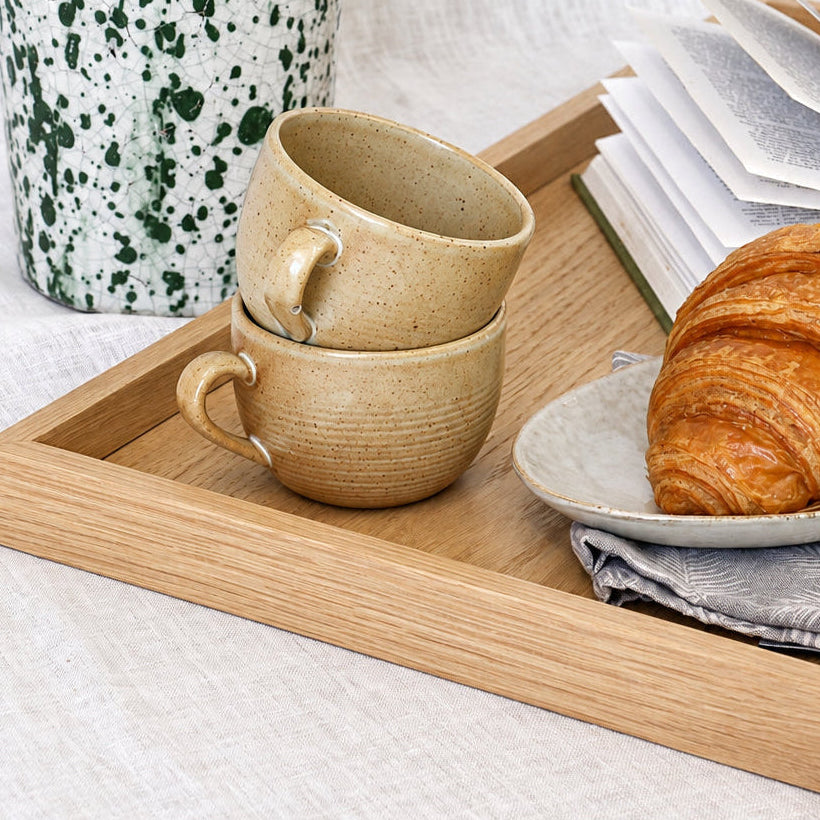 A styled breakfast scene on a light wood tray with two stacked beige ceramic mugs, an open book, a croissant on a small plate, and a tall white speckled planter with green leaves against a white linen backdrop.