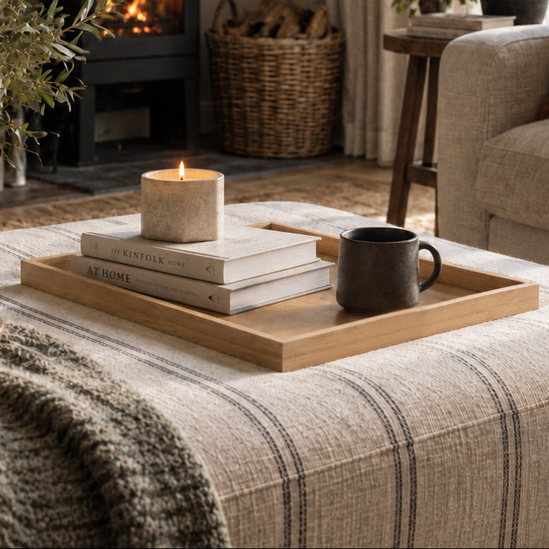 A cozy living room scene with a black wood-burning stove, beige sofa, striped throw blanket, and a wooden tray holding stacked books and a candle on the ottoman.