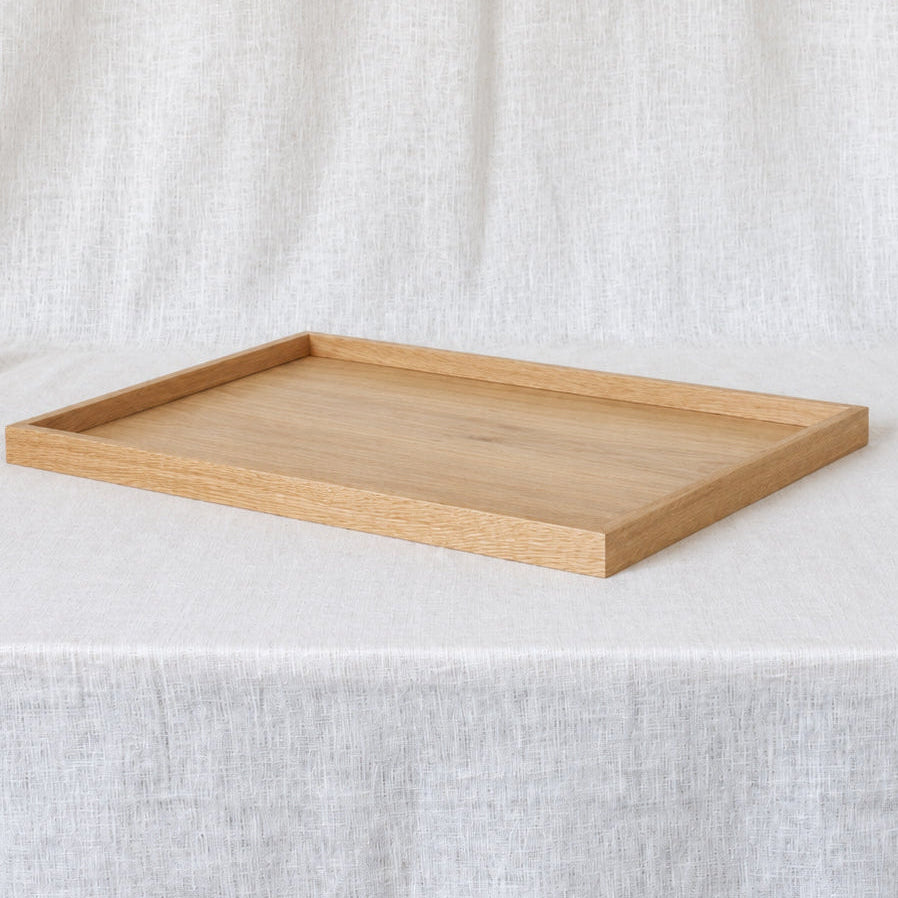 A rectangular light oak wooden tray with a raised rim, displayed on a draped white linen fabric background.