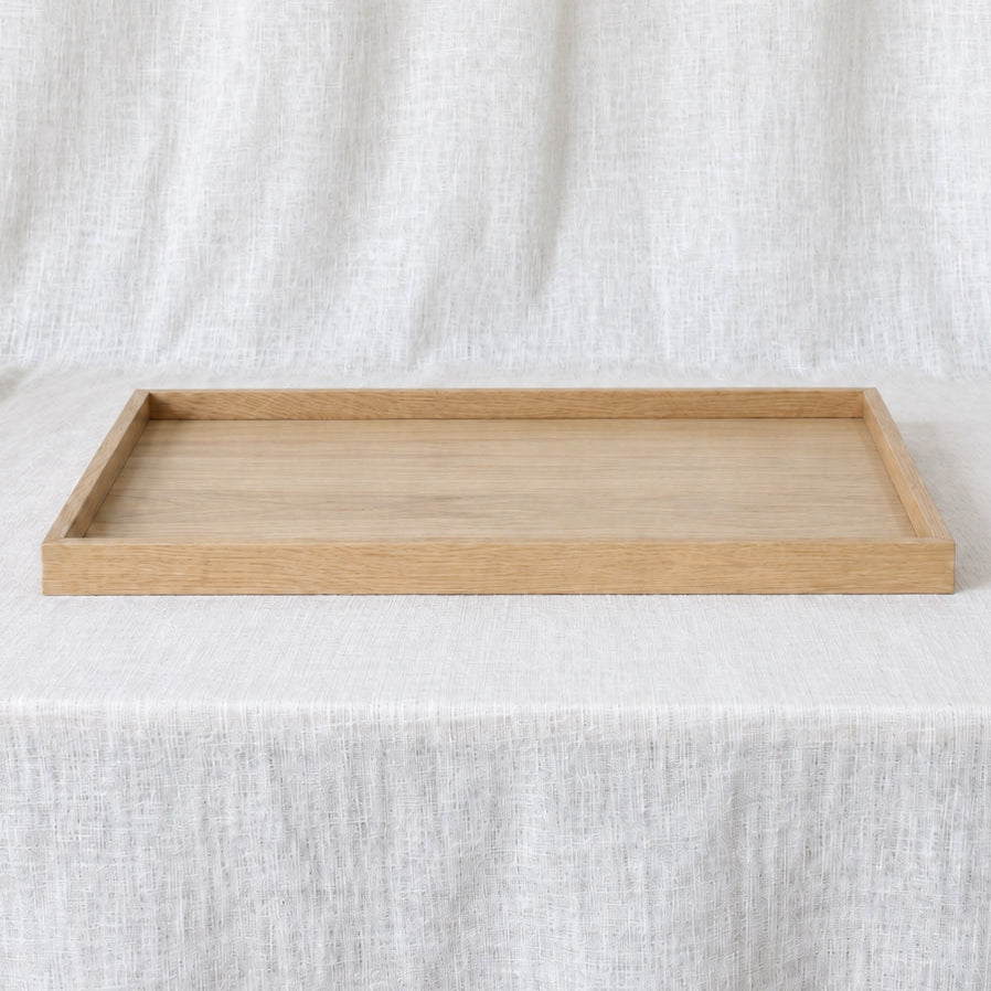 A rectangular light oak wood tray with a natural finish, displayed on a draped white linen fabric background.
