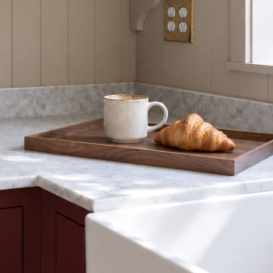 A styled kitchen corner with cream paneled walls, open floating shelves holding bowls, plates, and plants, a white marble countertop, dark red cabinets, and a window with natural light. A walnut tray on the counter holds a white mug and a croissant.
