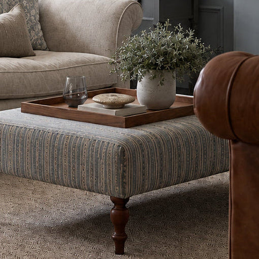 A styled interior room scene viewed through open paneled doors, showing a beige sofa, striped ottoman, side table with lamp, fireplace, and a brown leather armchair in a blue-gray living room.