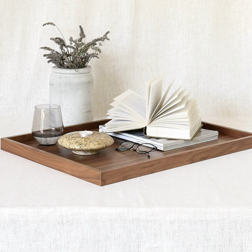 A rectangular wooden serving tray styled with an open book, a small vase of dried stems, a glass, and a small dish on a draped white fabric background.
