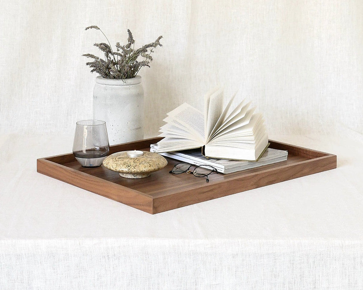 A rectangular wooden serving tray styled with an open book, a small vase of dried stems, a glass, and a small dish on a draped white fabric background.