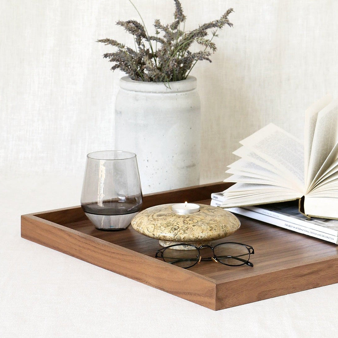 A rectangular wooden serving tray styled with an open book, a small glass, a round stone candle holder, and a pair of eyeglasses, set against a neutral white linen backdrop with a ceramic vase of dried lavender stems.