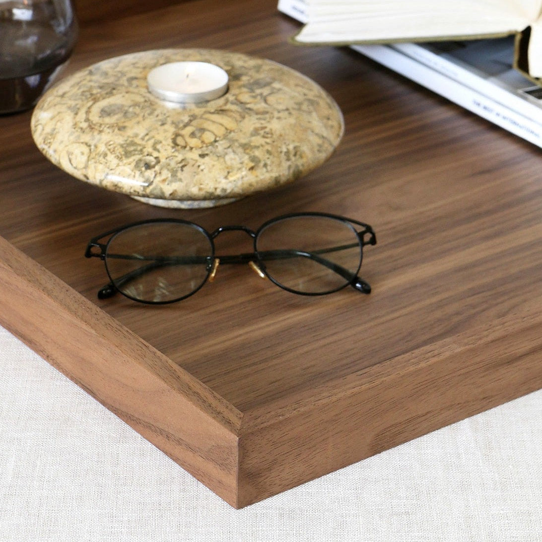 A rectangular wooden tray with straight corners sits on a light fabric surface, styled with an open book, a folded magazine, a small round stone holder with a candle, and a dark glass cup.