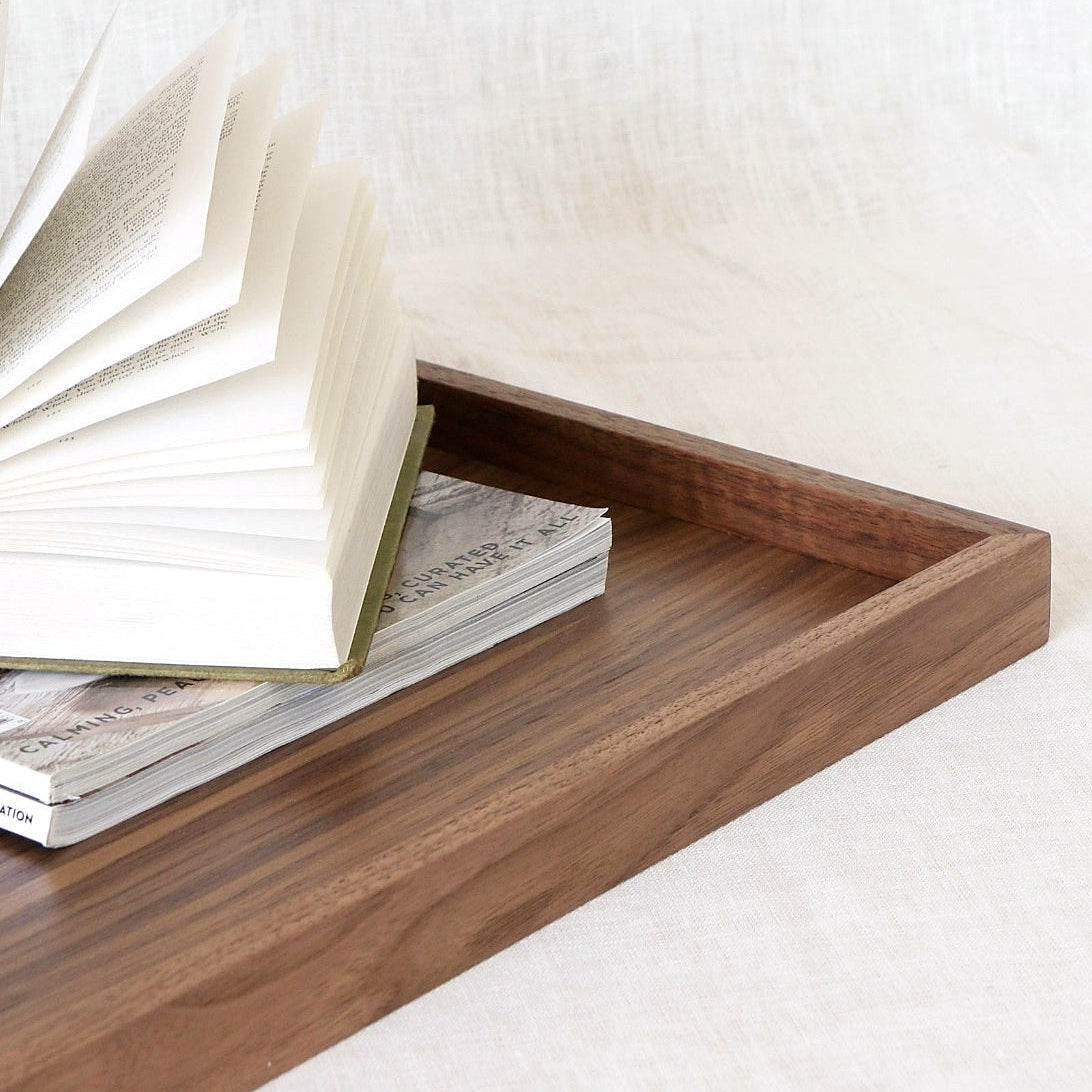 A rectangular wooden tray with a warm medium-brown finish, styled with an open book and a magazine on a light beige textured fabric surface.