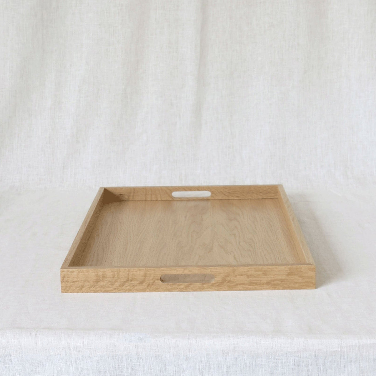 A rectangular light oak wood serving tray with two cutout handles, displayed on a draped white fabric background.