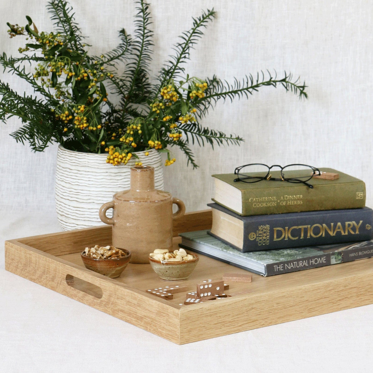 A light natural oak serving tray with cutout handles, styled with a small ceramic vase of yellow flowers, a small brown jug, and a stack of books on a white tablecloth against a white linen background.