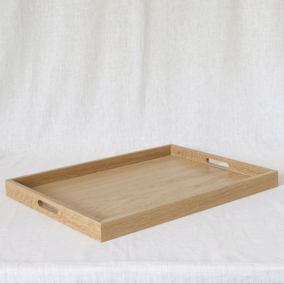 A rectangular light oak wooden serving tray with two cutout handles on the sides, displayed on a draped white fabric background.