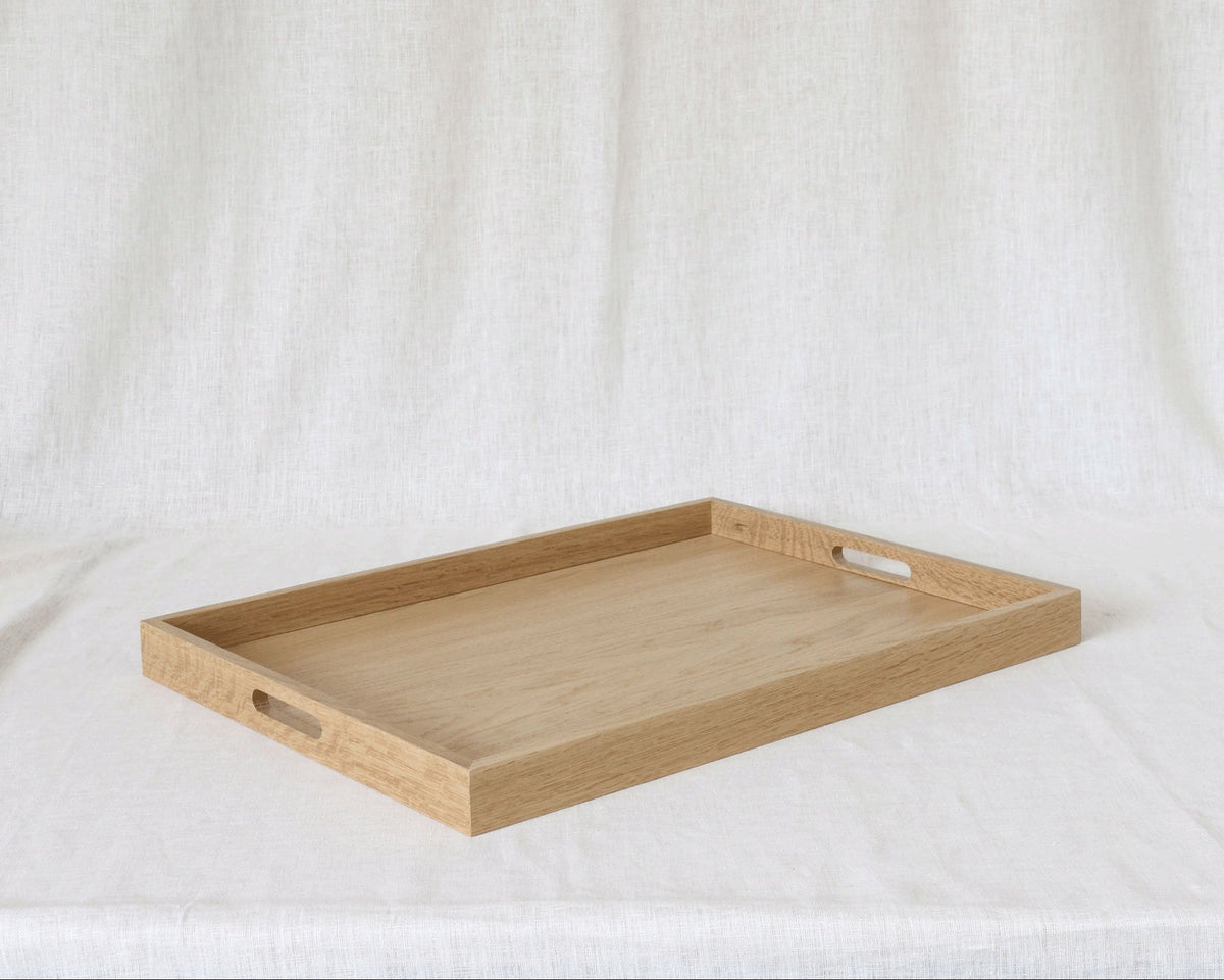 A rectangular light oak wooden serving tray with two cutout handles on the sides, displayed on a draped white fabric background.