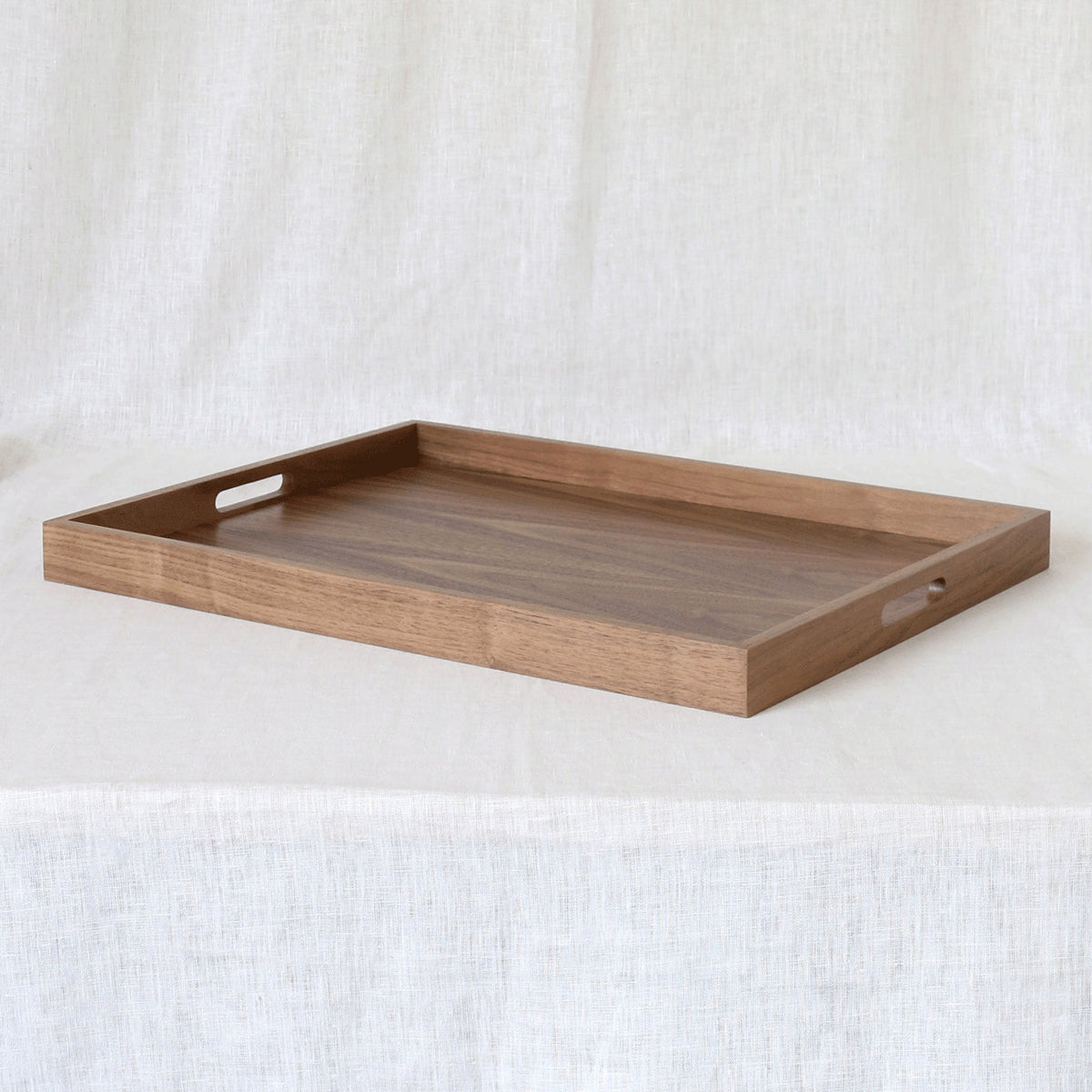 A rectangular wooden serving tray with two cutout handles on the sides, displayed on a draped white linen fabric background.
