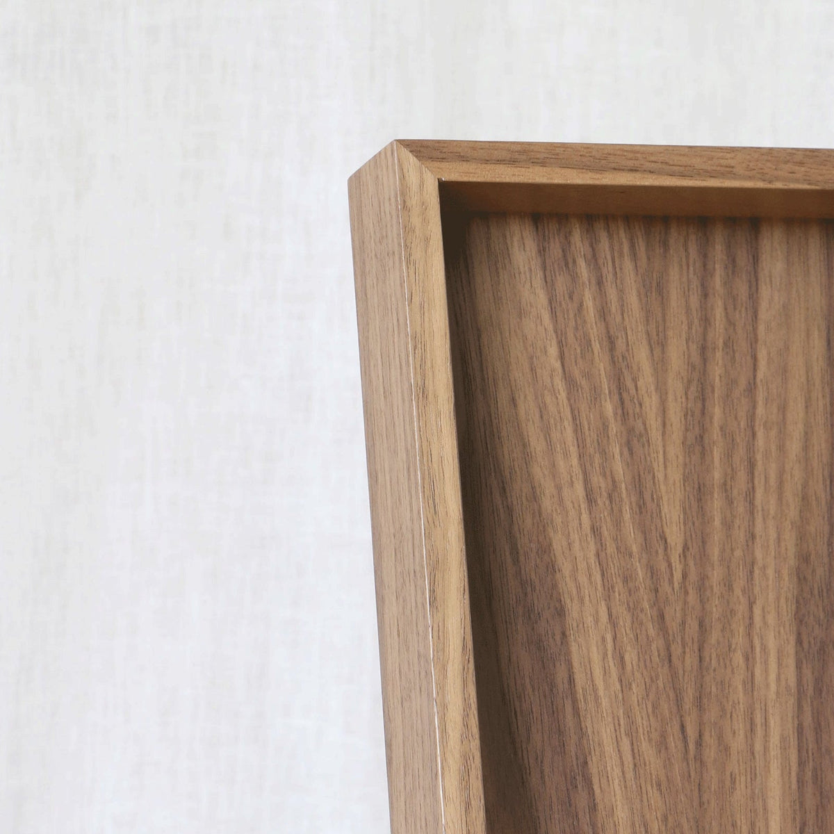 Close-up of a wooden tray frame corner with a warm medium-brown wood grain against a light beige textured wall.