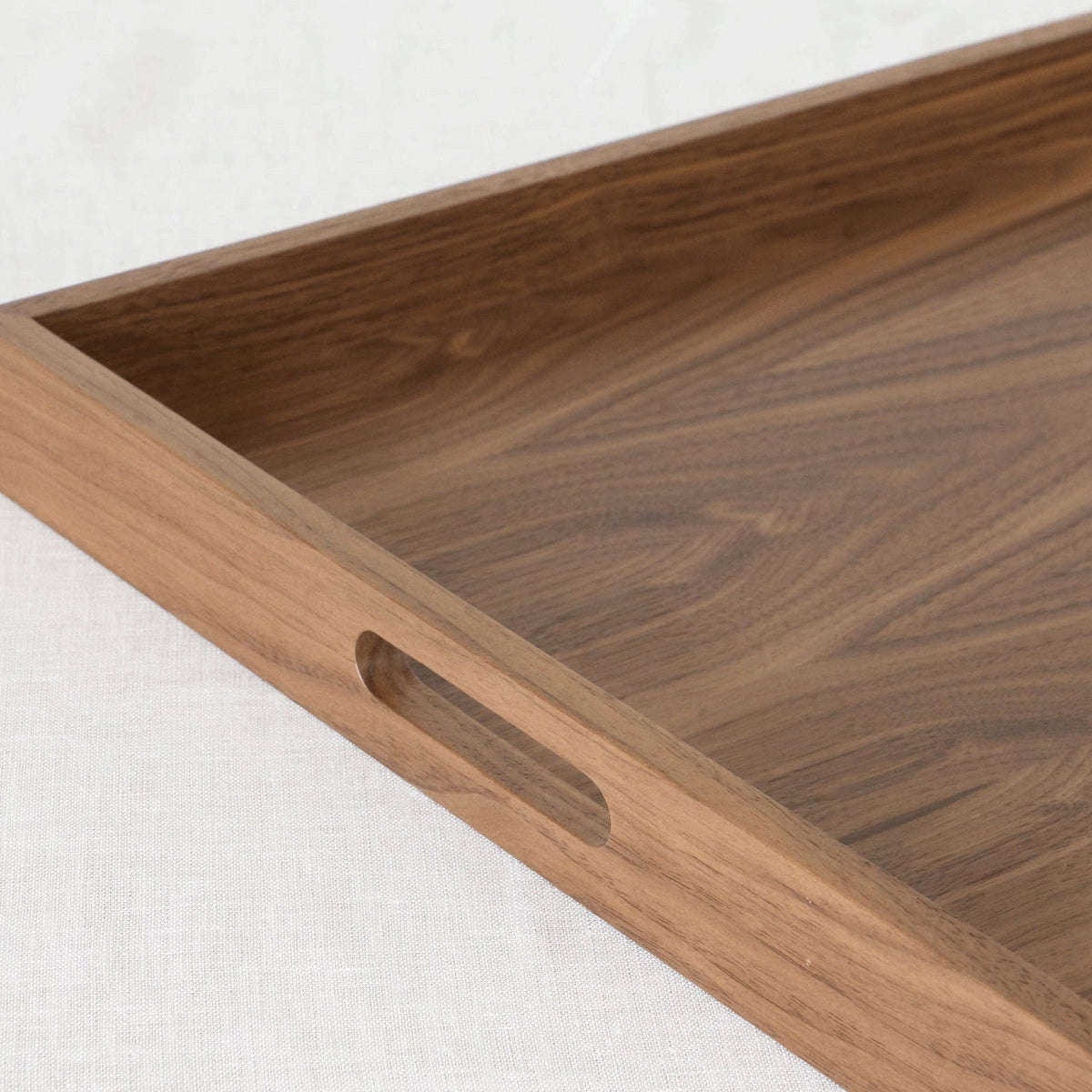 A close-up of a rectangular wooden serving tray with a recessed handle cutout on one side, shown on a light textured fabric background.