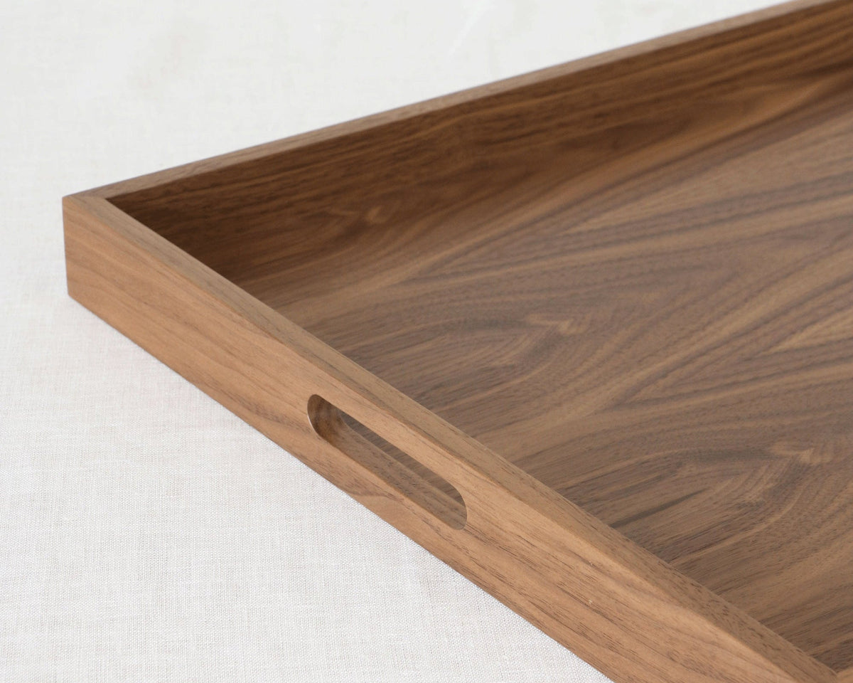 A close-up of a rectangular wooden serving tray with a recessed handle cutout on one side, shown on a light textured fabric background.