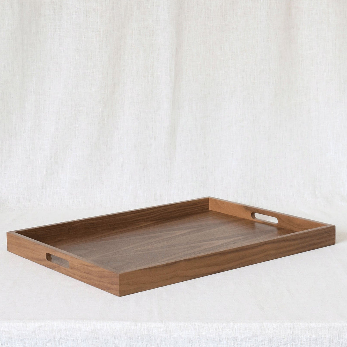A rectangular wooden serving tray with two cutout handles, resting on a draped white fabric background.