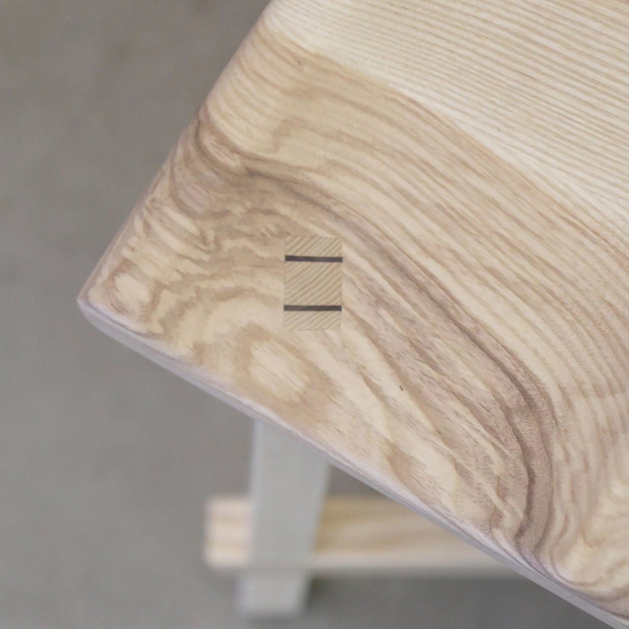Close-up of a wooden stool with a carved seat joinery detail on a grey floor.