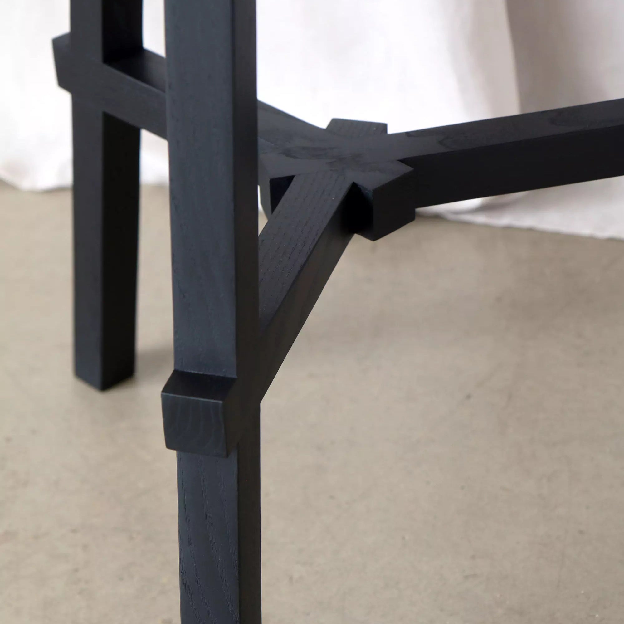 Close-up of a black wooden chair leg on a beige floor with a white curtain in the background.