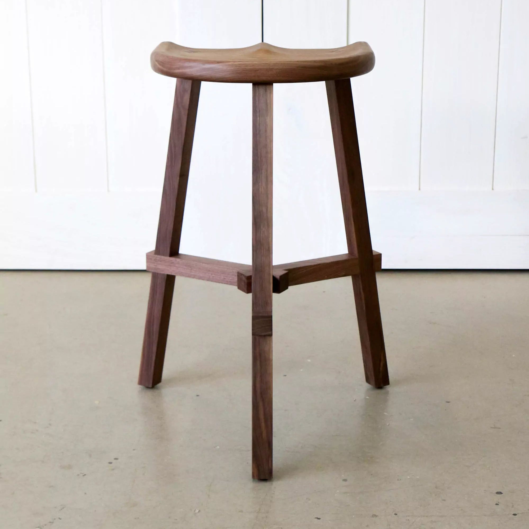Walnut stool on a light-colored floor with a white panelled wall in the background
