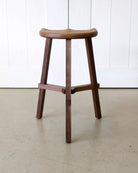 Walnut stool on a light-colored floor with a white panelled wall in the background