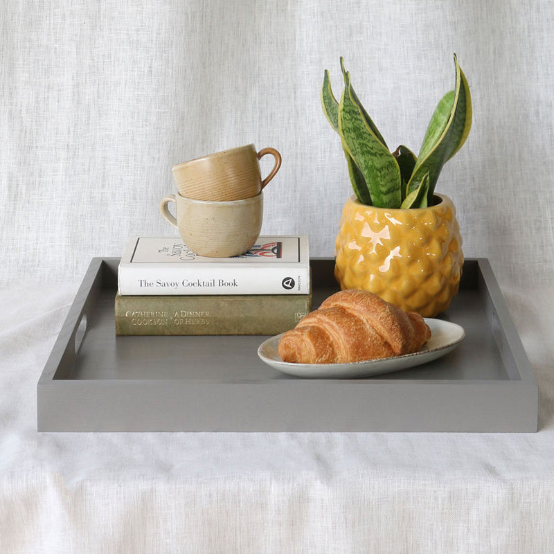 Grey ottoman tray featuring a yellow pineapple-style planter, croissant, stacked mugs, and books.