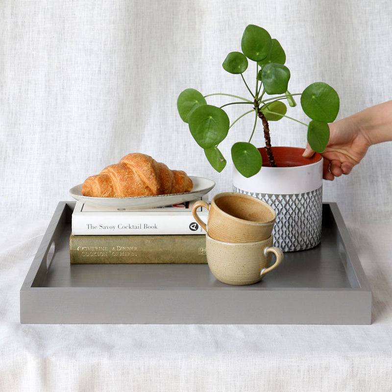 Grey ottoman tray styled with a plant, croissant, ceramic mugs, and books, with a hand placing the plant.