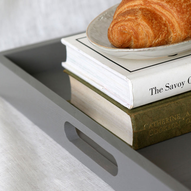Grey ottoman tray styled with a croissant on a plate and stacked books including “The Savoy Cocktail Book.”