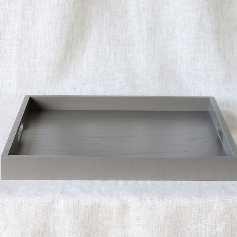 Front-facing view of a rectangular grey ottoman tray with handles on each side, set against a light fabric background.
