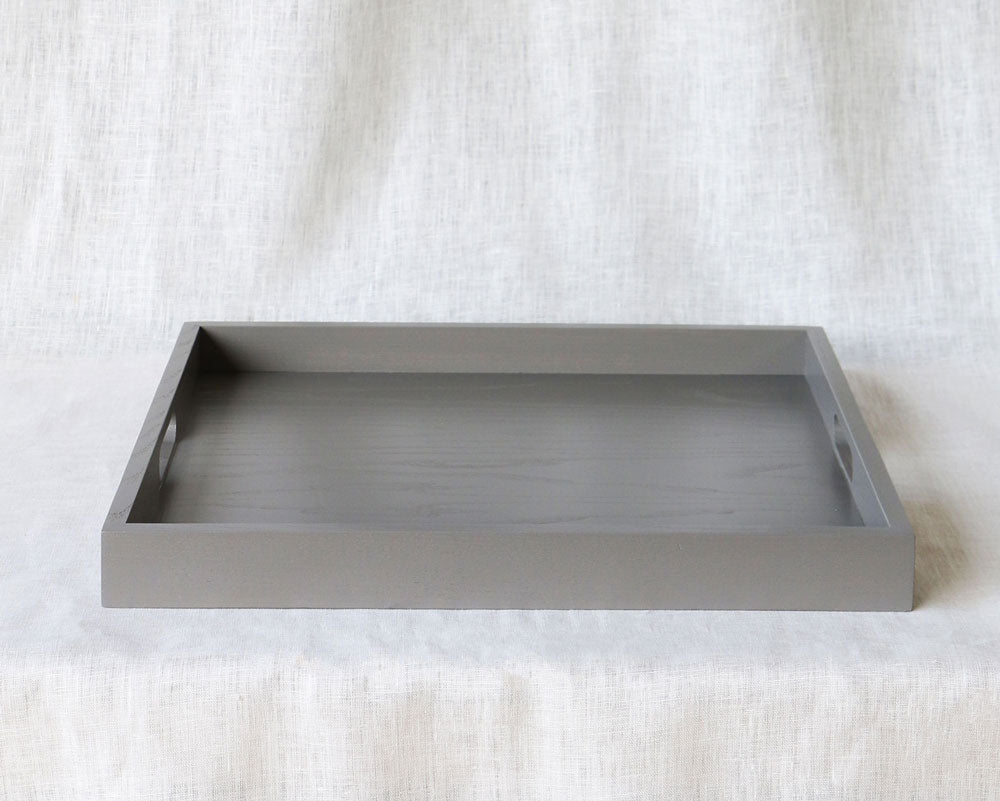 Front-facing view of a rectangular grey ottoman tray with handles on each side, set against a light fabric background.